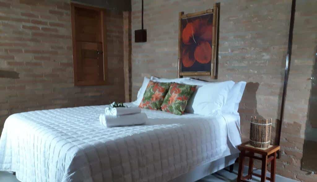 Quarto acolhedor da Vila das Mangabeiras com parede de tijolos, quadro colorido e cama confortável com roupa de cama clara e almofadas florais. Essa é uma das dicas de onde ficar em Corumbau.