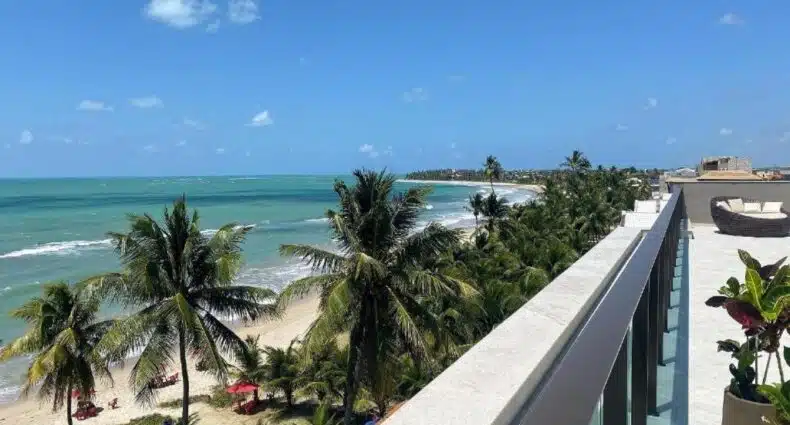 Vista da Praia dos Carneiros a partir do Flat Studio beira mar com piscina privativa com coqueiros, céu azul e mar de tonalidades esverdeadas e azuis, vista da varanda de um edifício. Ilustrando uma das opções de onde ficar em Tamandaré.