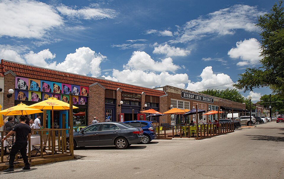 Loja e restaurante no Bichop Arts District em Dallas movimentado com fachada colorida, carros estacionados na rua e pessoas aproveitando as mesas do lado de fora sob guarda-sóis.