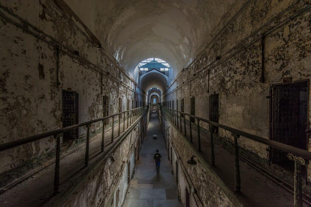 Corredor escuro com celas prisionais em ambos os lados e uma figura caminhando ao fundo, na Eastern State Penitentiary, para representar o que fazer na Filadélfia.