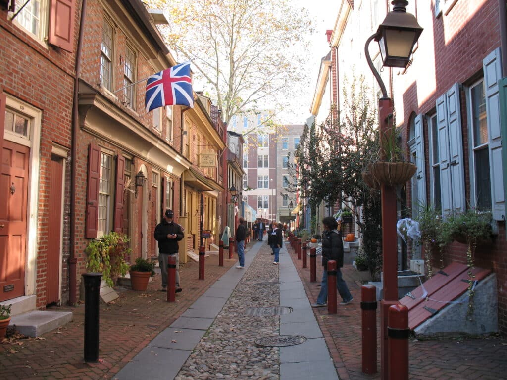 Rua comercial em centro histórico com bandeira do Reino Unido, prédios de tijolos, lanternas de rua e pessoas caminhando na calçada, em Elfreth’s Alley, para representar o que fazer na Filadélfia.