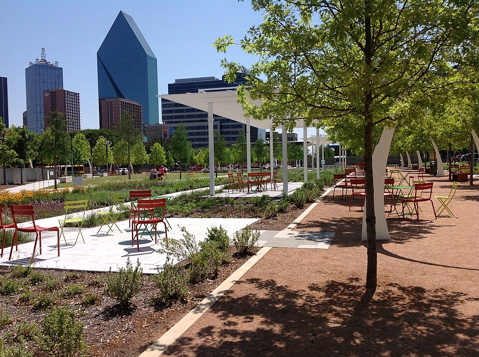 Parque urbano Klyde Warren Park em Dallas com prédios arranha-céus e árvores ao fundo, mesas e cadeiras vermelhas espalhadas pelo espaço ajardinado.