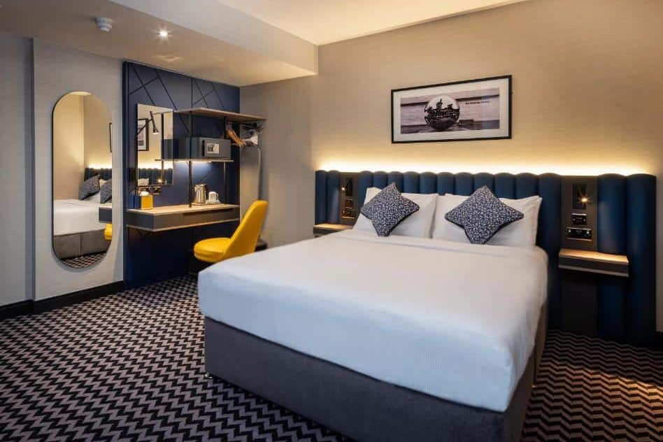 Quarto do Academy Plaza Hotel com cama de casal, cabeceira acolchoada azul, luminária de parede e espelho circular na parede. Cerca de madeira preta decorativa atrás da cabeceira. Piso com padrão geométrico em preto e branco. A foto faz parte do post de onde ficar em Dublin.