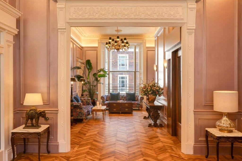 Luxuosa sala de estar no Albany House com piso de madeira em espinha de peixe, móveis clássicos floridos, plantas grandes e lustres dourados em ambiente opulento de estilo vitoriano.