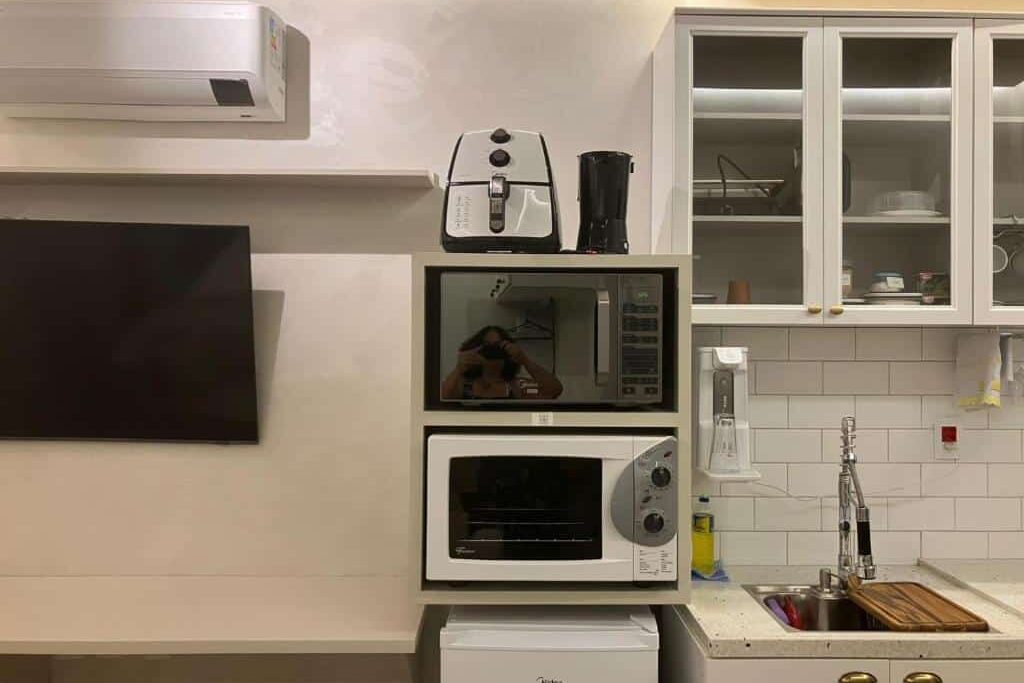 Cozinha compacta no Apart-hotel Theotókos Flats com eletrodomésticos modernos, incluindo microondas, torradeira e cafeteira em bancada branca. Prateleiras de vidro acima da pia exibem talheres e utensílios. Televisão montada na parede.