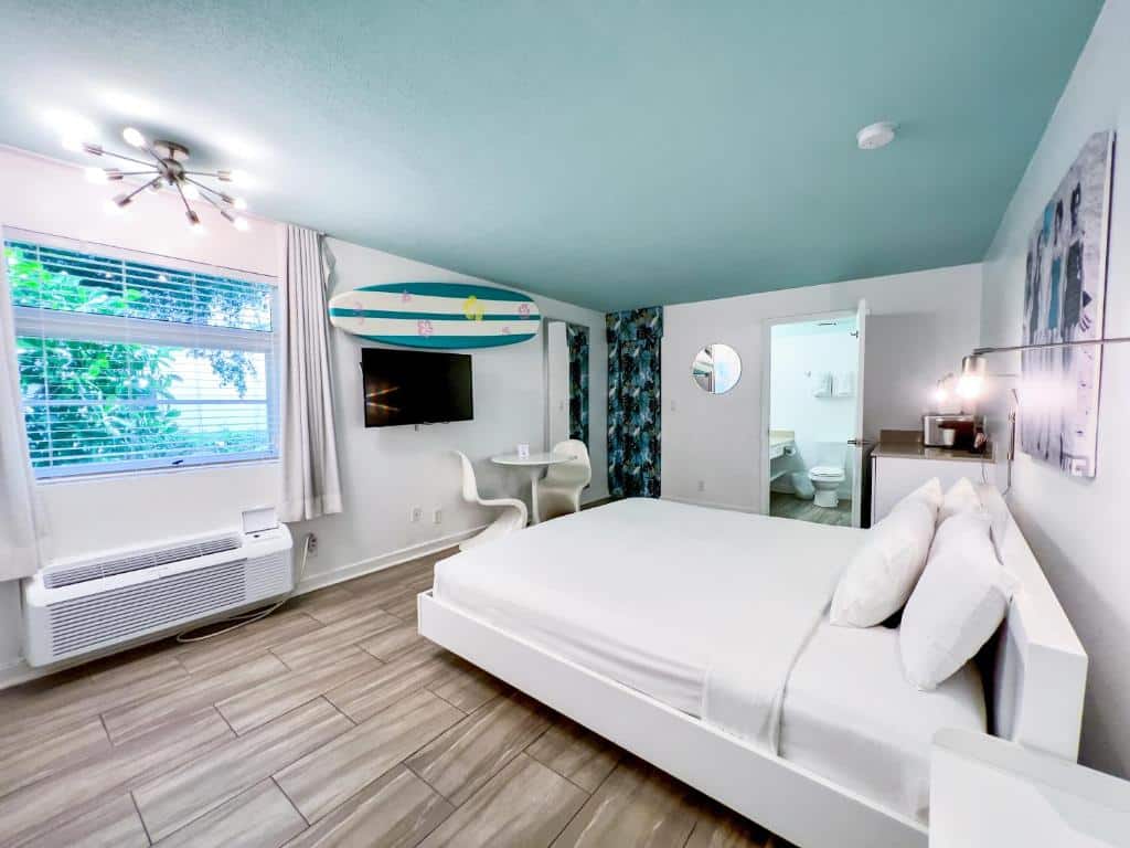 Quarto acolhedor do Aqua Hotel com paredes brancas, piso de madeira clara e decoração de praia com prancha de surfe na parede. A imagem mostra uma cama de casal branca com travesseiros e uma mesa redonda com cadeiras ao lado da janela.