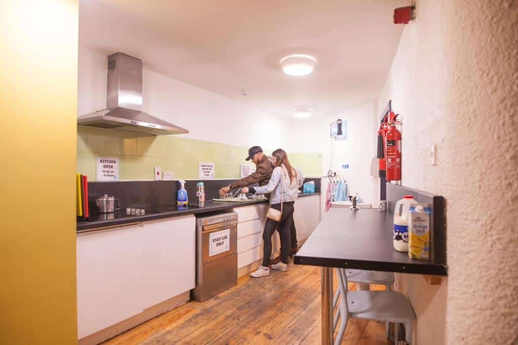 Casal jovem preparando refeição juntos em cozinha no Ashfield Hostel com piso de madeira e eletrodomésticos modernos.