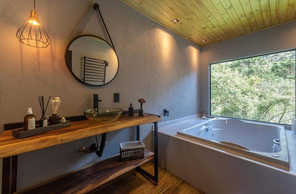Banheiro rústico com piso de madeira, parede clara, iluminação natural, pia de madeira e cuba de vidro, espelho redondo, torneira preta e banheira de hidromassagem com vista da natureza.