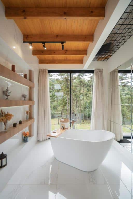 Banheiro moderno com banheira branca oval, prateleiras em madeira clara, cortinas claras e vista da natureza através da ampla janela.