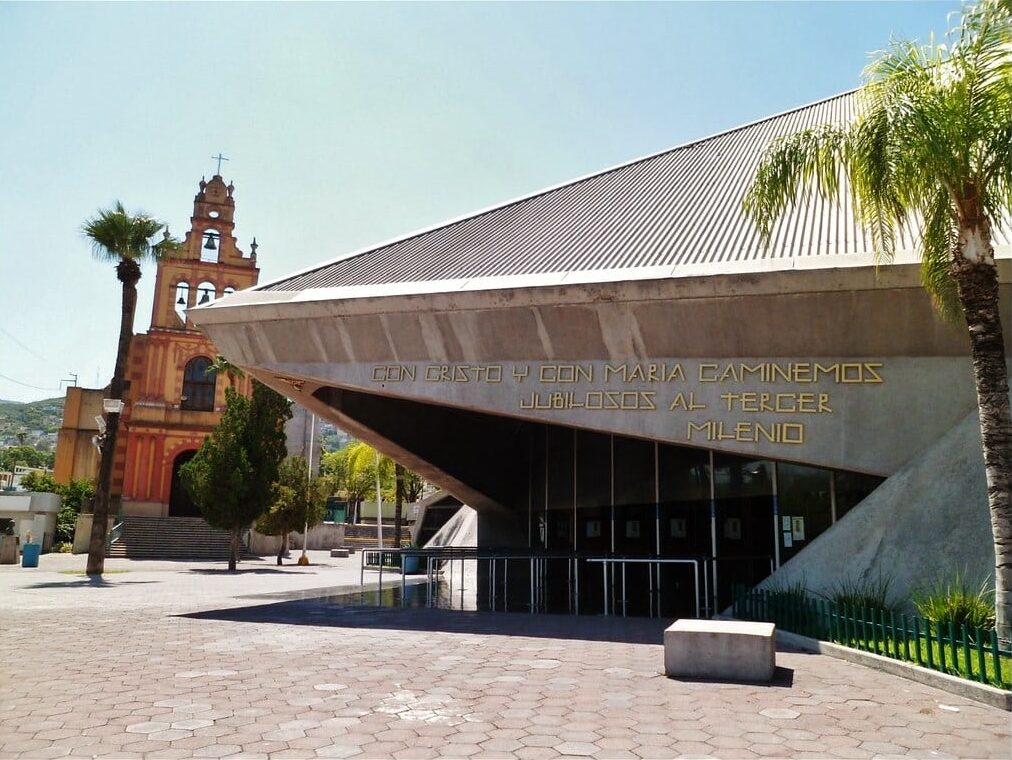 Edifício moderno com telhado metálico ondulado e igreja histórica ao fundo, em ambiente urbano com palmeiras. Essa é a Basílica de Guadalupe.