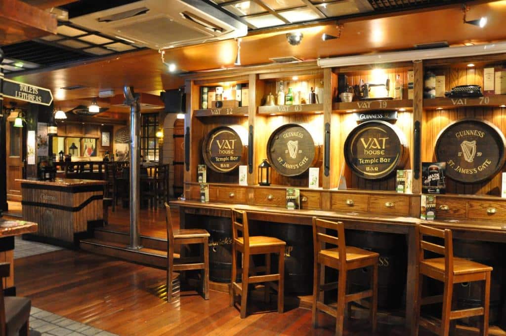 Um interior rústico e aconchegante de um bar irlandês tradicional no Blooms Hotel. Mesas de madeira, cadeiras e prateleiras decoradas com barris e garrafas de Guinness, criando uma atmosfera típica de um pub. O ambiente é iluminado por luzes quentes.