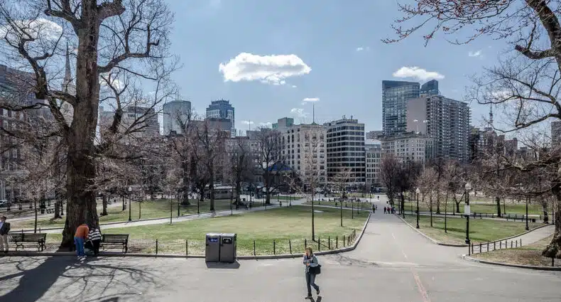 Parque urbano, o Boston Common, com edifícios modernos ao fundo, pessoas caminhando e bancos em um dia ensolarado com nuvens no céu. Imagem para ilustrar post sobre o que fazer em Boston.