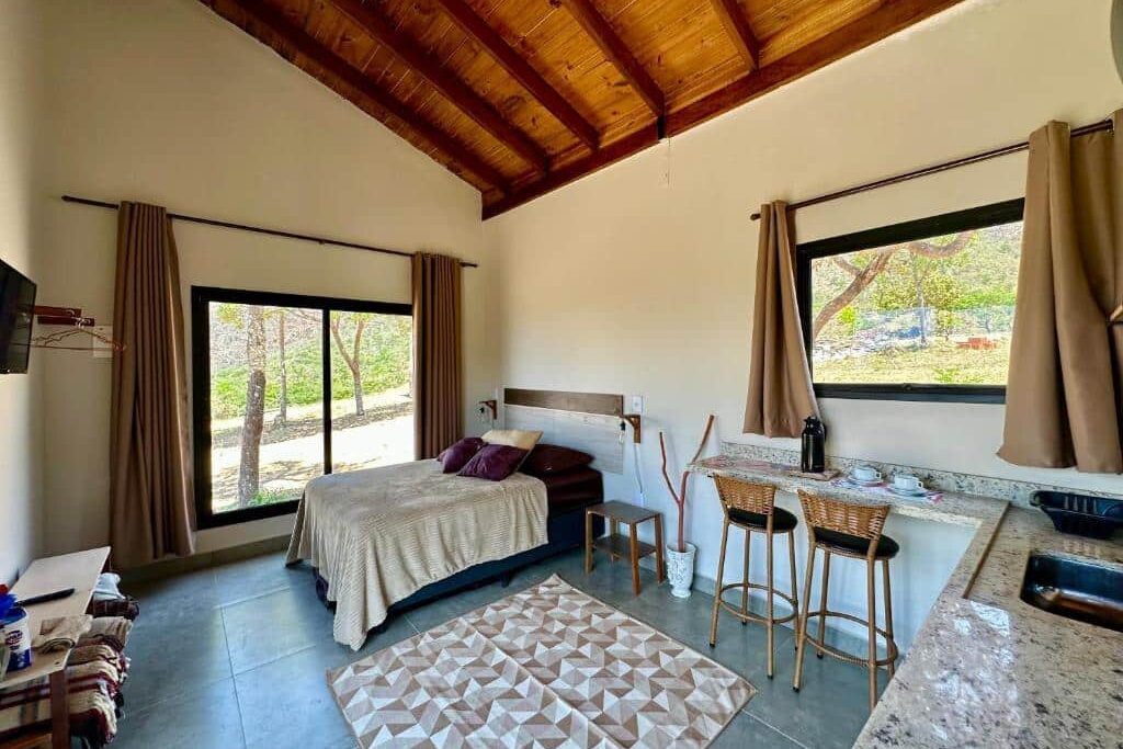 Quarto rústico dos chalés canário de minas com teto de madeira, amplas janelas e móveis de design minimalista. Cama confortável com lençóis claros, acompanhada de mesa de jantar com duas cadeiras e aparador de madeira escura.