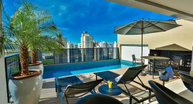 Piscina com borda elevada em cobertura de prédio residencial, com guarda-sol e cadeiras de praia ao redor, cercada por arranha-céus no Quality Hotel Flamboyant. Representa pousadas em Goiânia.