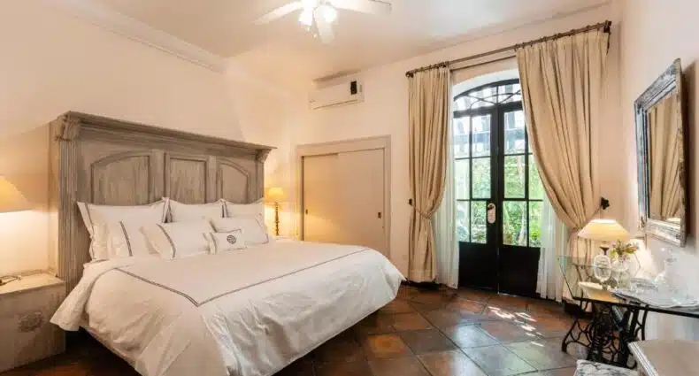 Quarto aconchegante do Villa Ganz Boutique Hotel com mobília de madeira rústica, cortinas bege e janela com vista para área externa arborizada. Representa hotéis em Guadalajara.
