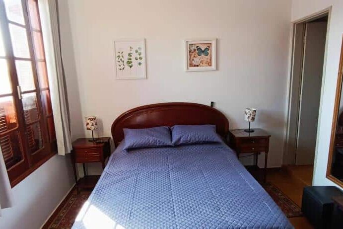 Quarto de casal na Casa Rosário Centro com cama de madeira escura, decorado com quadros na parede e lâmpada de cabeceira.