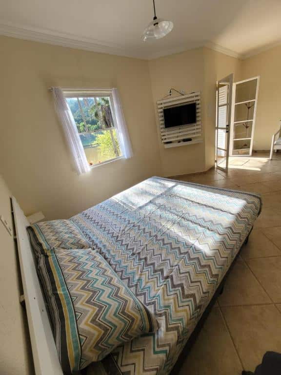 Quarto ensolarado do Chalés Sítio Dois Lagos com janela dando vista para jardim, cama de casal com edredom estampado azul, marrom e bege, TV na parede e armário branco. Ilustra onde ficar em Amparo.