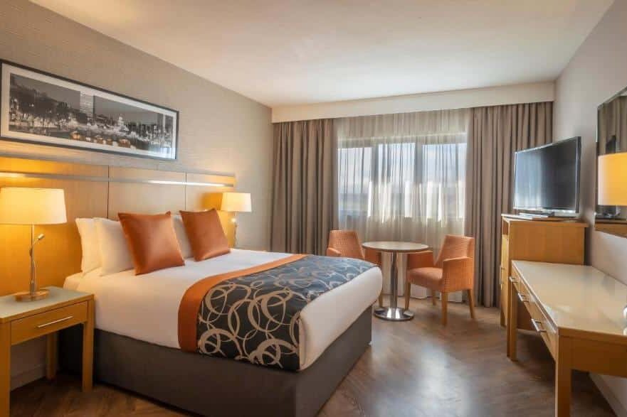 Quarto do Clayton Hotel Ballsbridge com cama de casal, decoração elegante em tons de laranja e bege, além de mesa de trabalho e cadeiras.