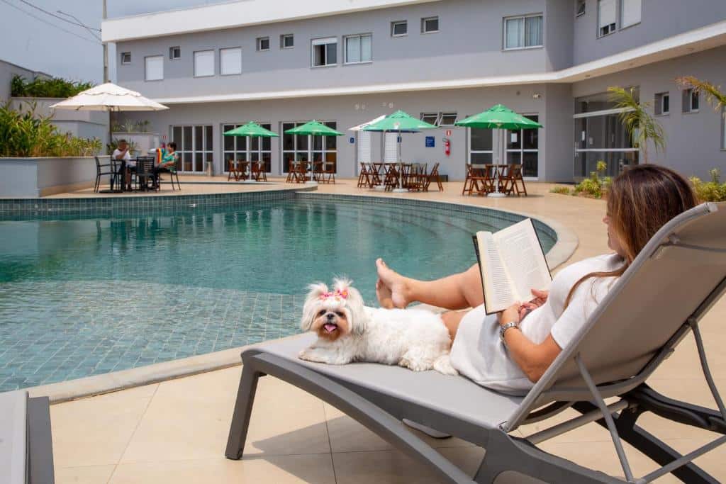 Uma mulher lendo um livro relaxadamente deitada em uma espreguiçadeira próxima a uma piscina, com uma cadela branca deitada a seu lado, em um hotel chamado Comfort Hotel Serra da Canastra com área de lazer ao ar livre repleta de guarda-sóis e mesas. Ilustra onde ficar na serra da canastra