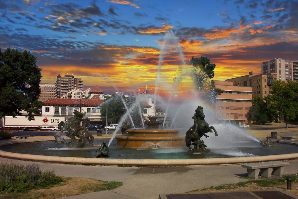 Praça central com bela fonte luminosa e estátuas, ao pôr do sol com céu incrível em tons de laranja e azul.