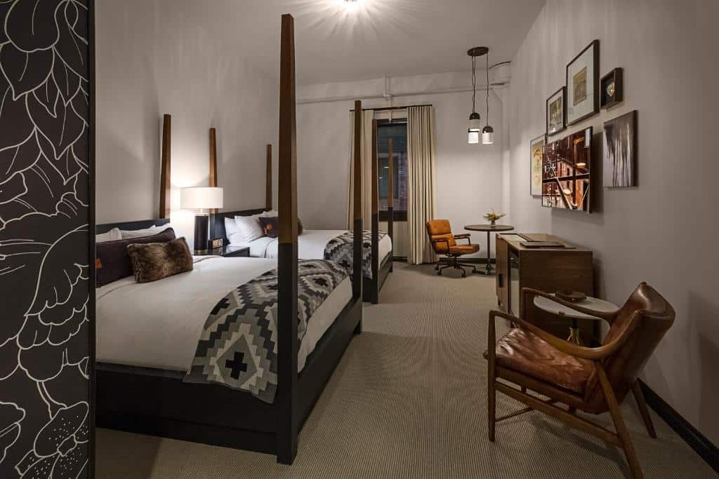 Quarto do Crossroads Hotel com cama de dossel de madeira, decoração neutra e tons de marrom, parede com design floral, mesa de escritório e cadeira de couro.