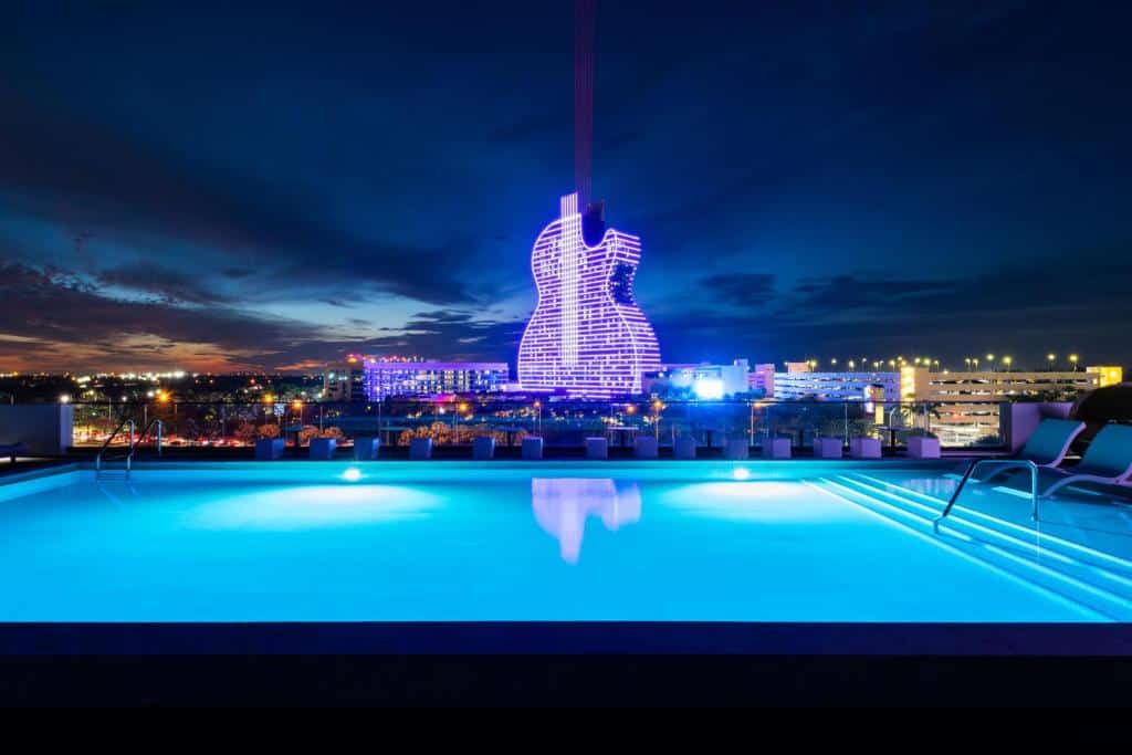 Edifício em forma de violão em frente ao Dolce by Wyndham Hollywood iluminado em cores azul e rosa na noite, com piscina refletindo a paisagem com vários prédios iluminados ao redor.