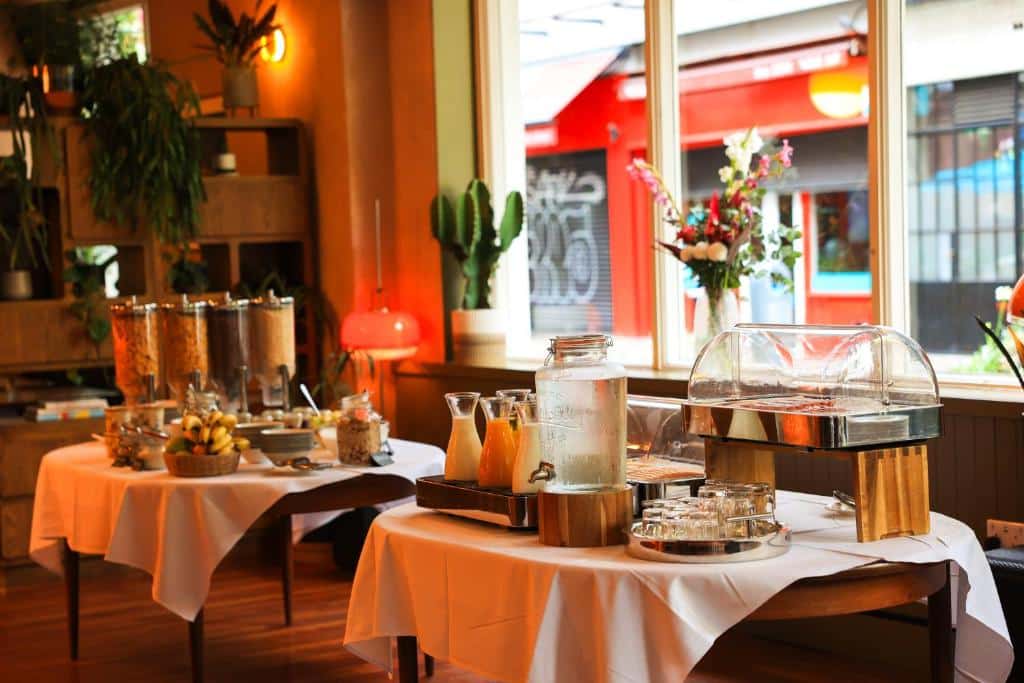 Mesa de refeição em restaurante no Drury Court Hotel com decoração rústica e plantas verdes, pronta para servir clientes com variedade de sucos, molhos e temperos sobre a toalha laranja.