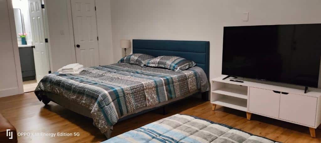 Quarto do Edercozy com cama de casal com roupa de cama listrada em tons de cinza, azul e branco, televisor grande em móvel branco, paredes claras e piso de madeira. Essa é uma das dicas de onde ficar perto do Hard Rock Stadium.