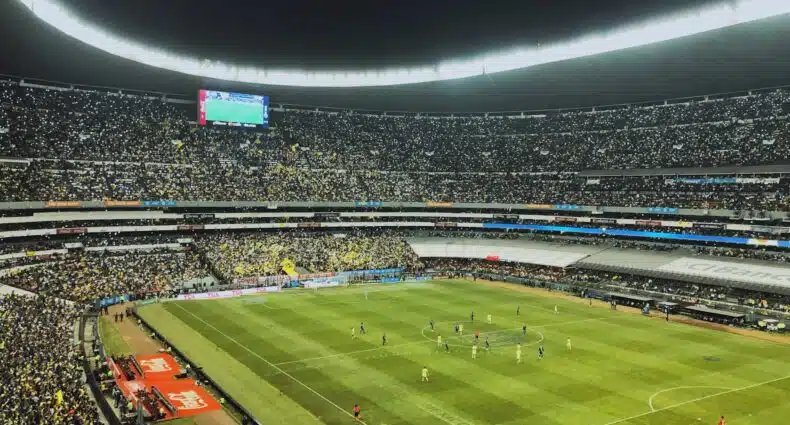 Estádio lotado em evento esportivo à noite com jogadores em campo e público nas arquibancadas. A imagem ilustra o post sobre hotéis perto do Estádio Azteca.