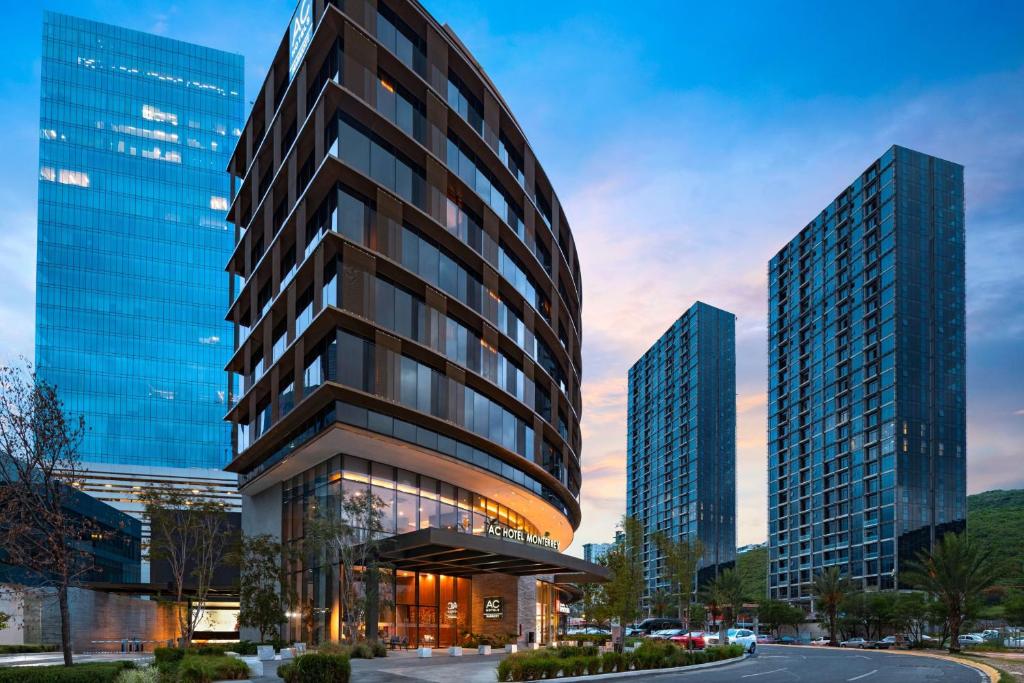 Imponente edifício de vidro e aço com entrada iluminada do hotel AC Hotel Monterrey, situado em distrito empresarial moderno com torres residenciais ao fundo.