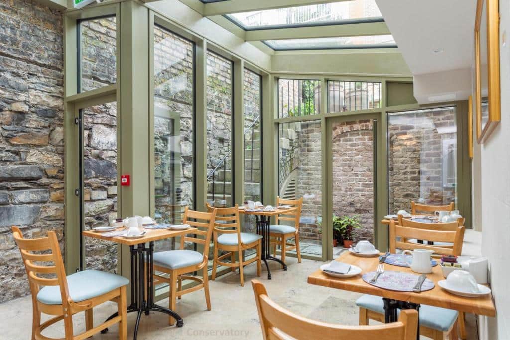 Área de jantar do Gardiner Lodge com paredes de pedra e teto de vidro, com mesas e cadeiras de madeira e uma decoração com plantas.