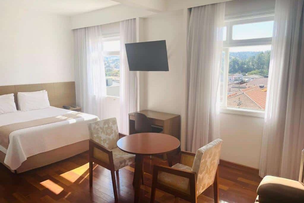 Quarto de Grande Hotel Amparo confortável com janela panorâmica, cama de casal, mesa redonda e cadeiras decorativas em ambiente aconchegante e iluminado. Ilustra onde ficar em Amparo.