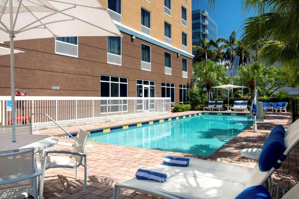 Piscina do Hampton Inn Hallandale Beach-Aventura com guarda-sóis brancos, espreguiçadeiras azuis e edificação de vários andares no fundo em ambiente tropical com palmeiras.