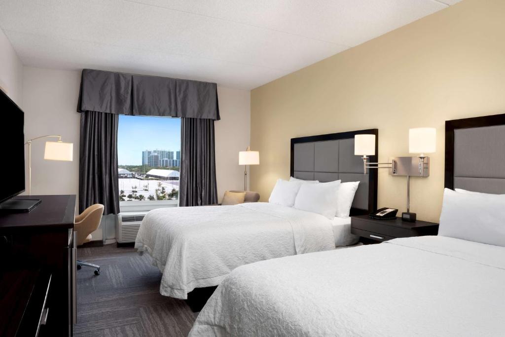 Moderno quarto do Hampton Inn Hallandale Beach-Aventura com duas camas de casal, móveis escuros e janela com vista para uma cidade com prédios altos. Essa é uma das dicas de onde ficar perto do Hard Rock Stadium.