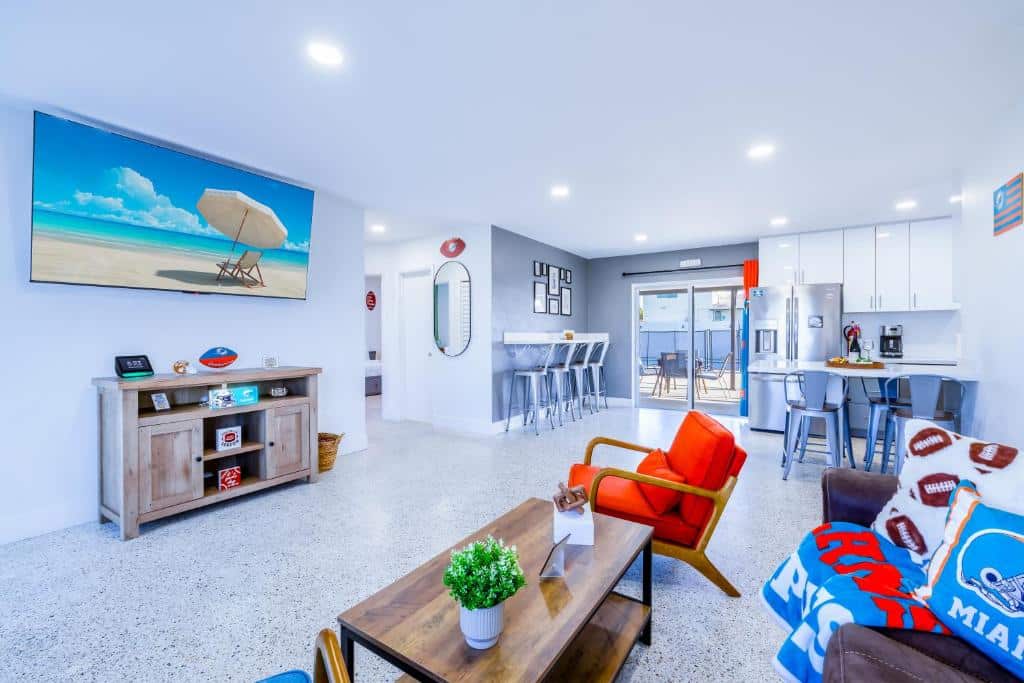 Sala de estar ampla em estilo moderno com decoração colorida e esportiva, incluindo móveis de madeira, cadeira de couro laranja, grande TV com imagem de praia ensolarada, e diversos objetos e acessórios relacionados a esportes no Havana Blue.