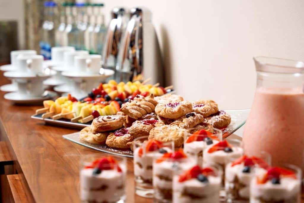 Lanches e frutas no café da manhã do Herbert Park Hotel and Park Residence, incluindo biscoitos, tortas e copos com bebida cremosa rosa, em um ambiente de cozinha acolhedor com prateleiras de vidro.