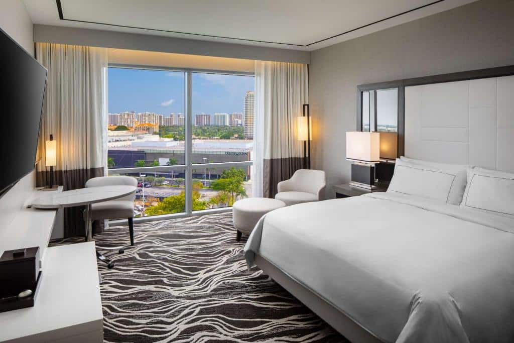 Quarto do Hilton Miami Aventura com vista panorâmica da cidade, com grandes janelas, cortinas bege e marrons, cama de casal branca, poltrona e mesa de trabalho. Essa é uma das dicas de onde ficar perto do Hard Rock Stadium.