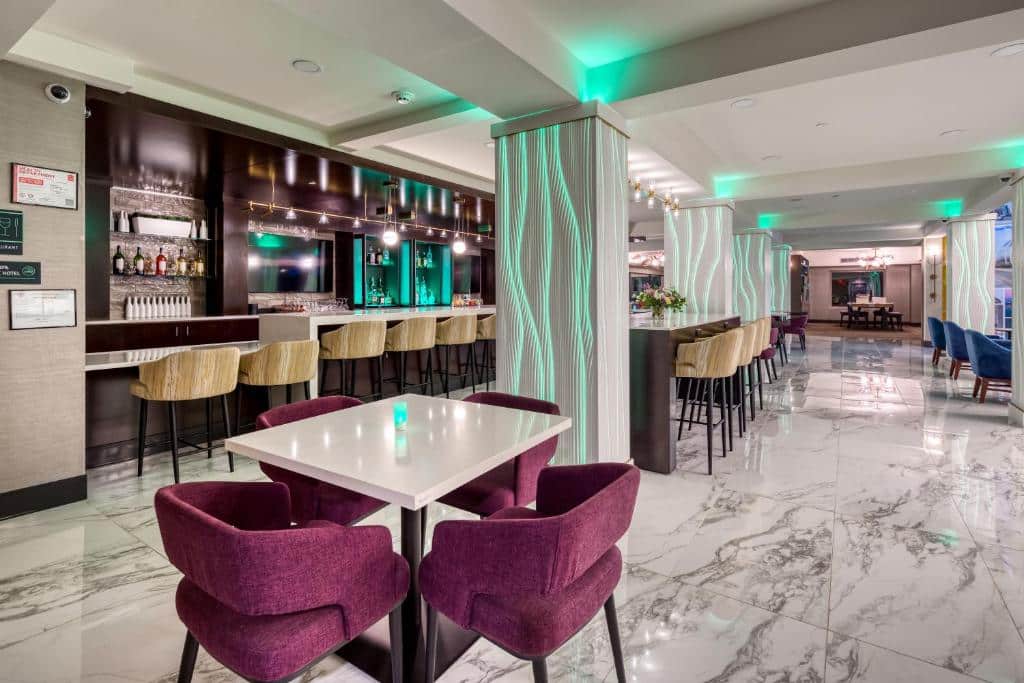 Moderna sala de bar do Holiday Inn Kansas City - Downtown com decoração elegante em tons escuros e iluminação verde, com balcão de madeira, cadeiras de veludo roxo e mesas brancas sobre piso de mármore.