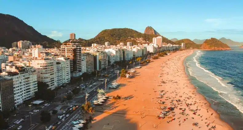 Praia de Copacabana, Rio de Janeiro, com montanhas e edifícios ao fundo, lotada de pessoas na areia em um dia ensolarado, para representar onde ficar em Copacabana.