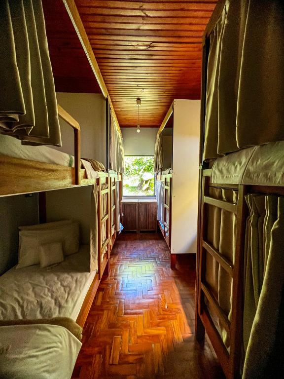 Corredor aconchegante de alojamento com beliches de madeira, roupa de cama bege e iluminação quente, com janelas dando vista para área externa