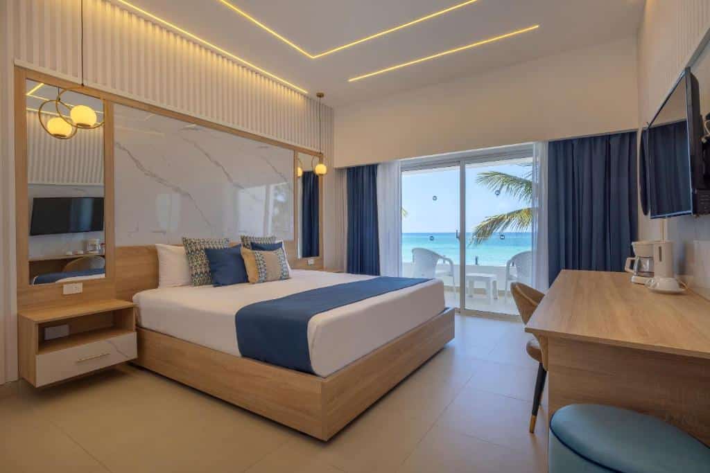 Quarto do Hotel HM Bavaro Beach, em Punta Cana, à beira-mar com cama de casal, móveis de madeira clara, janela panorâmica com vista para a praia e palmeiras.