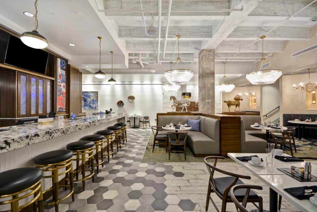 Salão de jantar elegante do Hotel Indigo Kansas City - The Crossroads com móveis de madeira e detalhes dourados, piso de ladrilhos hexagonais e lustres de cristal pendentes do teto alto.