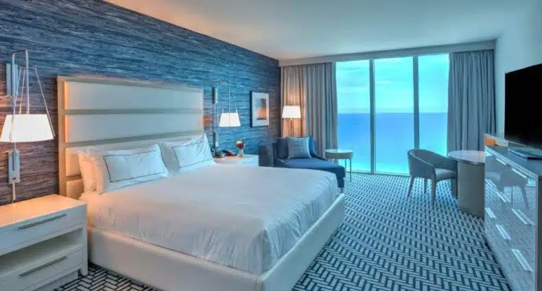 Quarto do Hotel Maren Fort Lauderdale com vista panorâmica do oceano azul durante o pôr do sol, com paredes revestidas de painel de madeira em tons escuros, cama de casal branca, poltrona azul e cortinas pesadas. Imagem para ilustrar post sobre onde ficar em Fort Lauderdale.