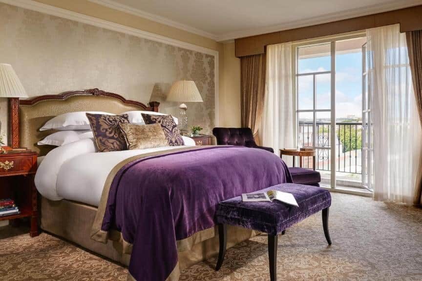 Quarto do InterContinental Dublin by IHG com cama de casal imponente, almofadas decorativas e cobertor roxo. Grandes janelas com cortinas claras e vista para um jardim externo. Mesa de canto com abajures e objetos decorativos, e banco acolchoado roxo próximo à cama.
