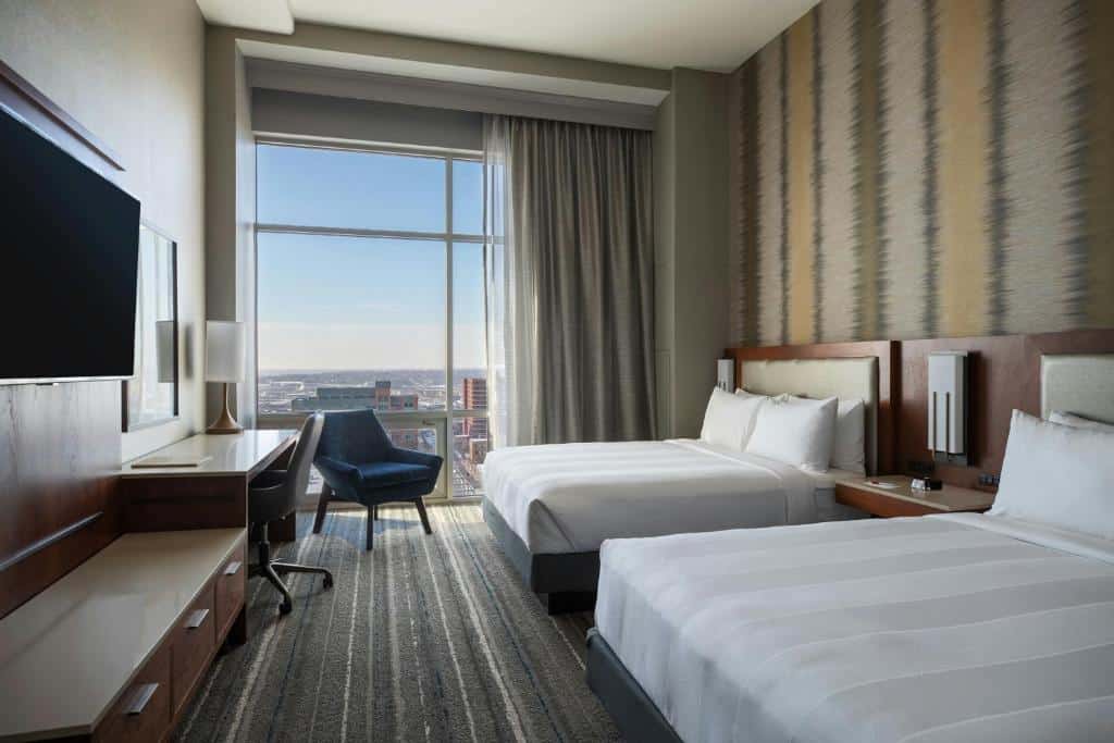 Quarto do Kansas City Marriott Downtown com vista panorâmica da cidade, decorado em tons neutros e equipado com duas camas de casal, TV de tela plana na parede e cadeira de escritório azul escura junto à janela.
