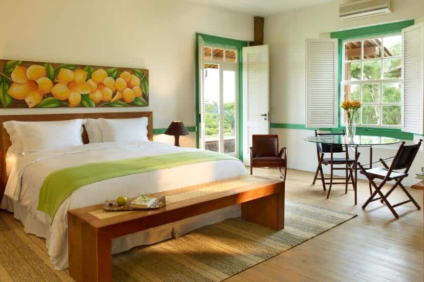 Quarto aconchegante do Lake Vilas Charm Hotel & SPA com cama de casal, obra de arte com flores amarelas, janela com vista para vegetação e mesa de jantar com cadeiras de madeira.