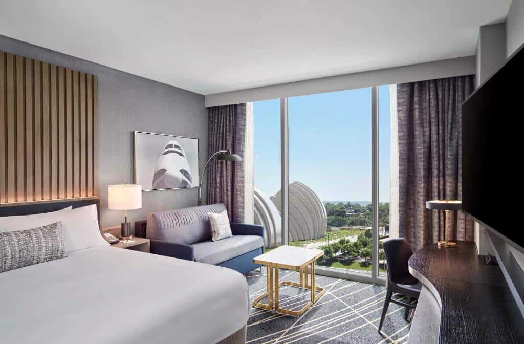Quarto do Loews Kansas City com vista panorâmica da cidade. Grandes janelas com cortinas cinza permitem entrada de luz natural. Sofá azul e móveis de madeira compõem o ambiente sofisticado. Obra de arte abstrata em tons neutros decora a parede. Atmosfera acolhedora e serena.