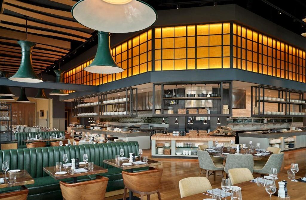 Restaurante do Loews Kansas City com design moderno e industrial, com mesas acolhedoras em tons de verde, iluminação pendente e painel de vidro refletindo a cozinha aberta.