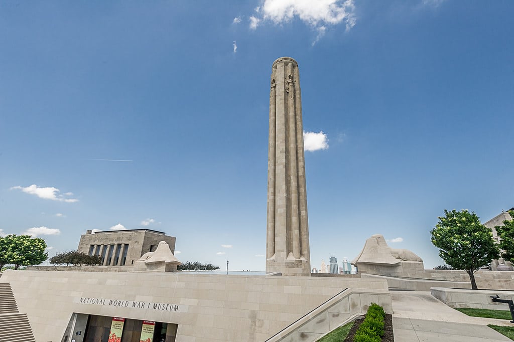 Torre comemorativa de concreto armado da Segunda Guerra Mundial com entrada destacada, em frente a um céu azul com nuvens brancas, cercada por áreas verdes. Essa é uma das dicas de o que fazer em Kansas City.