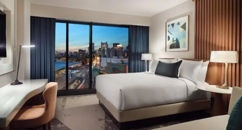 Quarto do Omni Boston Hotel com vista panorâmica da skyline ao anoitecer, com edifícios iluminados e luzes do tráfego abaixo. Imagem para ilustrar post sobre onde ficar em Boston.